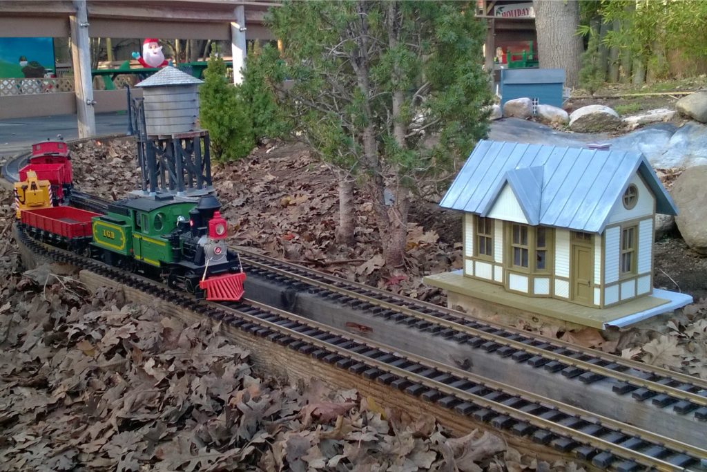 Model train approaches station passing water tank and small grove of pine trees.